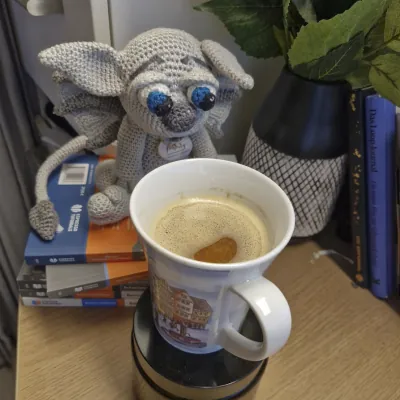 Gideon sitzt auf einem Buchstapel und schaut direkt in eine Kaffeetasse